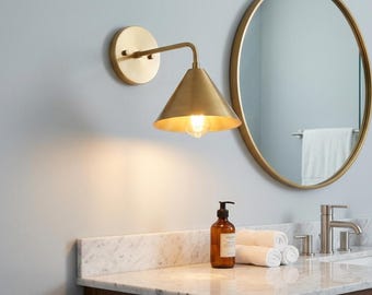 May include: A brass wall sconce with a conical shade and a round backplate is mounted on a light grey wall. A round gold-framed mirror and a marble countertop with a soap dispenser and rolled towels are also visible.