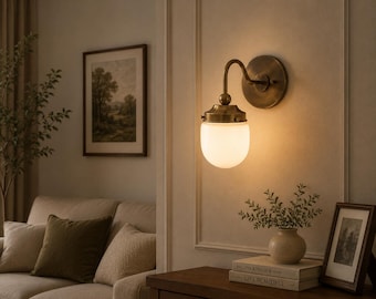 May include: A vintage-style wall sconce with a brass finish and a frosted glass shade. The sconce is mounted on a cream-colored wall, casting a warm glow. The image also shows a framed landscape painting, a sofa, and decorative items.