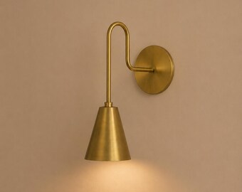 May include: A brass wall light with a conical lampshade and a curved arm. The light has a circular backplate and a warm glow. The fixture is made of a gold-toned metal. The overall design is minimalist.