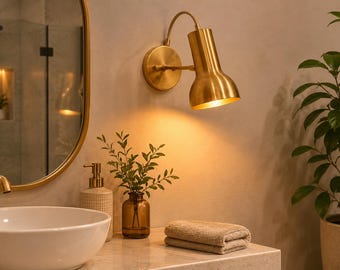 May include: A bathroom interior with a gold-coloured wall lamp, a round mirror, and a white sink. A small brown glass bottle with greenery and a folded beige towel are on the countertop. A potted plant is on the right.