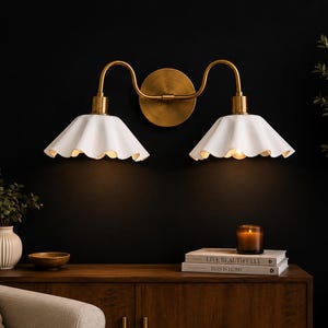 May include: A double-light wall sconce with a brass finish and two white, scalloped-edge shades. The fixture is mounted on a dark wall, with a warm glow emanating from the lights. The sconce is part of a home decor theme.
