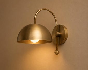 May include: A brass wall sconce featuring a dome-shaped shade and a curved arm. The sconce has a circular backplate and a small, spherical detail. The light bulb is visible inside the shade, casting a warm glow.