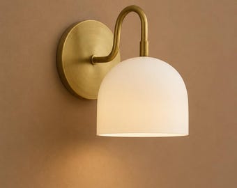 May include: A wall-mounted light fitting with a brass finish. It has a circular base, a curved arm, and a white, dome-shaped shade. The fitting is designed to provide ambient lighting.