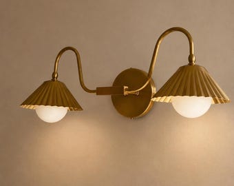 May include: A double wall light fixture in a brass finish. It features two lamps with pleated shades and spherical, white glass bulbs. The sconce is mounted on a round backplate, suitable for a bedroom or hallway.