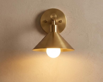 May include: A brass-colored wall sconce with a conical shade and a round backplate. The sconce features a single light bulb and is mounted on a neutral-colored wall. The design is minimalist and modern, suitable for various interior styles.