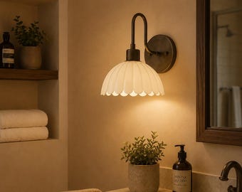 May include: A bathroom wall light with a white, scalloped lampshade and dark metal arm and base. The light illuminates a bathroom setting, with a mirror, sink, and potted plants. The fixture adds a touch of vintage charm to the space.