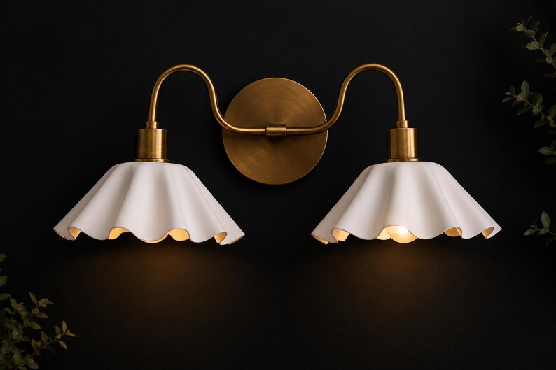 May include: A double-light wall sconce with a brass finish. The fixture features two white, scalloped-edge shades and a circular backplate. The sconce is mounted on a dark wall, with soft lighting emanating from the shades.