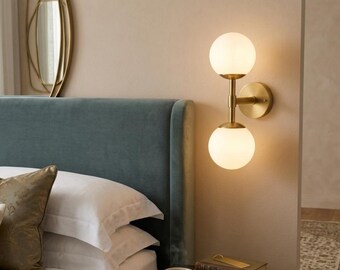 May include: A contemporary wall light featuring two frosted glass globe shades. The fixture has a brass finish and is mounted on a beige wall. The light is next to a teal headboard and a bedside table with books and a teacup.