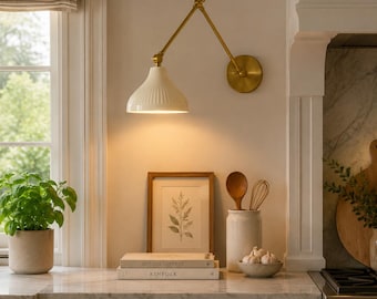 May include: A kitchen scene featuring a white and gold wall-mounted lamp with an adjustable arm. A framed botanical print, cookbooks, and a ceramic utensil holder with wooden spoons and a whisk sit on the countertop. A potted herb and a bowl of garlic are also visible.