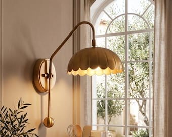 May include: A brass wall light with a scalloped lampshade. The light has a curved arm and a circular base. The fixture is mounted on a wall near a window with a view of trees and a neutral-toned curtain.
