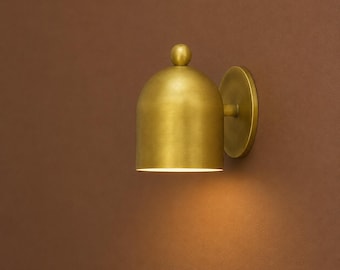 May include: A gold-coloured wall sconce featuring a bell-shaped shade and a small, spherical finial. The sconce has a circular backplate and emits a warm light. The fixture is mounted on a brown wall, creating a contrast with the gold finish.