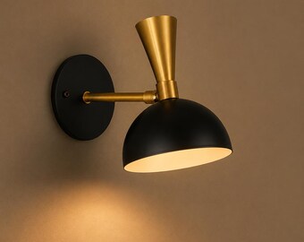 May include: A wall-mounted lamp featuring a circular black base, a horizontal gold arm, and a conical gold accent piece connecting to a matte black dome-shaped shade. The interior of the shade is white, and the lamp emits a warm, soft light.