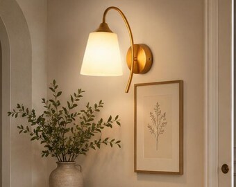 May include: A gold-finished wall sconce with a white glass shade. The sconce features a curved arm and a circular backplate. A framed botanical print and a vase with greenery are also visible in the image.