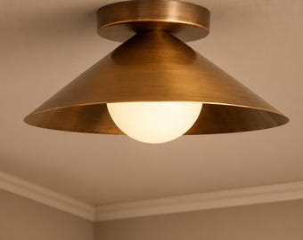 Puede incluir: Una lámpara de techo de montaje empotrado con una pantalla cónica ancha con acabado en latón antiguo cepillado. Una bombilla esférica de cristal blanco está centrada bajo la pantalla de metal, proporcionando una luz suave y difusa.