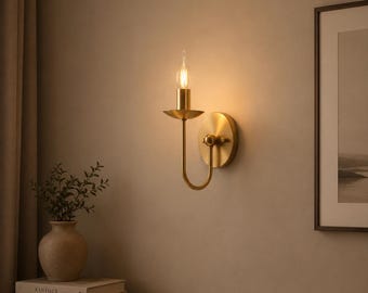 May include: A brass wall sconce with a candle-style light bulb. The sconce features a circular backplate and a curved arm. The light is lit, casting a warm glow. The sconce is fixed to a beige wall.