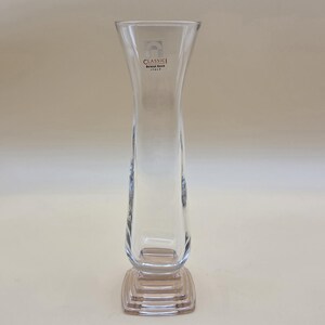 May include: A clear glass vase with a flared rim and a square, stepped base. The glass features the printed text "CLASSIC Bormioli Rocco ITALY" near the top. The vase has a slender, curved body that transitions into a solid, tiered square pedestal.