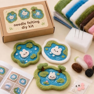 Puede incluir: Un kit de manualidades de fieltro con aguja que incluye una caja de cartón con una etiqueta que muestra tres escenas de estanques con conejitos blancos. Contiene lana de colores, un bloque de espuma blanco con tres agujas y un manual.