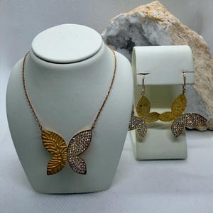 May include: A gold-toned butterfly necklace and matching earrings set. The butterfly wings are detailed with a textured pattern and encrusted with sparkling crystals. The jewelry is displayed on white stands against a neutral background.