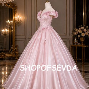 Könnte beinhalten: Ein bodenlanges, hellrosa Ballkleid mit schulterfreiem Ausschnitt, der mit Stoffblumen verziert ist, einem eng anliegenden Oberteil mit Blumenapplikationen und einem voluminösen Rock in Satinoptik. Der Text SHOPOFSEVDA ist sichtbar.