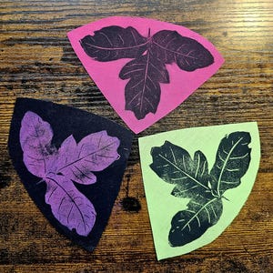 May include: Three triangular fabric patches featuring a dark leaf print. One patch is bright pink with a dark purple leaf, one is black with a light purple leaf, and one is light green with a dark green leaf, each showing detailed leaf veins.