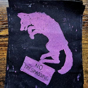 Puede incluir: Un parche de tela negra con un gráfico serigrafiado en color morado claro. El diseño muestra la silueta de un cánido saltando sobre un letrero rectangular que contiene el texto NO TRESPASSING en una fuente negrita sin remates.