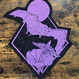 May include: A black and purple fabric patch featuring a bat in flight against a full moon, positioned above a moth resting on a flower. The design is enclosed within a diamond-shaped border, showcasing detailed line work in a vibrant purple hue.