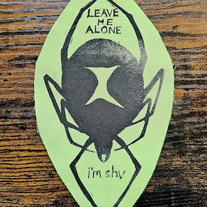 May include: A leaf-shaped fabric patch in light green featuring a black screen-printed graphic of a spider. The text LEAVE ME ALONE is printed above the spider body, and the text i'm shy is printed below the spider body in black ink.