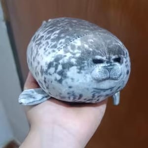 May include: A soft seal toy in grey and white, held in a hand. The seal has a rounded body, flippers, and a detailed face with small, dark eyes and whiskers. The toy is designed to be soft and cuddly, ideal for children.