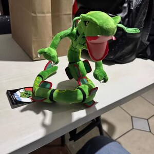 May include: A green plush toy of a serpentine creature with a red mouth and accents. The toy has a long, winding body with red and yellow details. A card with a picture of the creature is next to the toy.