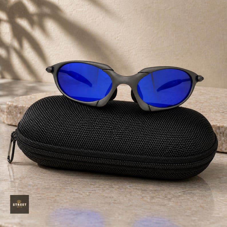 Peut inclure: Des lunettes de soleil grises avec des verres bleus reposent sur un &eacute;tui noir zipp&eacute;. Les lunettes de soleil ont un design sportif et moderne. L'&eacute;tui est rectangulaire et a une surface textur&eacute;e. Le fond est beige neutre.