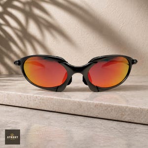 Peut inclure: Lunettes de soleil noires avec des verres rouge-orange. Les lunettes ont un design &eacute;l&eacute;gant et moderne avec une monture incurv&eacute;e et un pont. Les verres pr&eacute;sentent un d&eacute;grad&eacute; de rouge et d'orange. Les lunettes sont pos&eacute;es sur une surface claire.