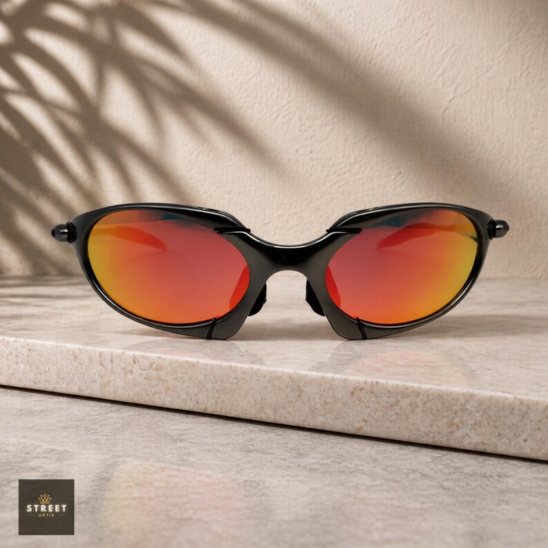 Peut inclure: Lunettes de soleil noires avec des verres rouge-orange. Les lunettes ont un design &eacute;l&eacute;gant et moderne avec une monture incurv&eacute;e et un pont. Les verres pr&eacute;sentent un d&eacute;grad&eacute; de rouge et d'orange. Les lunettes sont pos&eacute;es sur une surface claire.