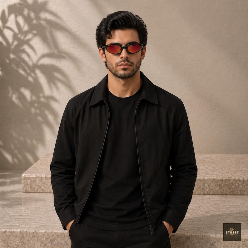 Peut inclure: Une personne portant une veste noire, un t-shirt noir et un pantalon noir, avec des lunettes de soleil teint&eacute;es de rouge. La veste a un col et une fermeture &eacute;clair. La personne se tient devant un fond de couleur neutre.