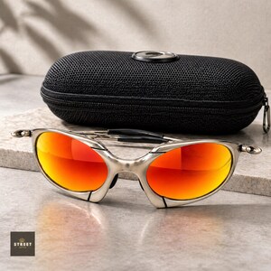 Peut inclure: Lunettes de soleil &agrave; monture argent&eacute;e avec des verres orange vif devant un &eacute;tui noir zipp&eacute;. Les lunettes ont un design moderne et &eacute;l&eacute;gant. Les verres refl&egrave;tent une teinte ardente. L'&eacute;tui a une surface textur&eacute;e et un d&eacute;tail argent&eacute;.
