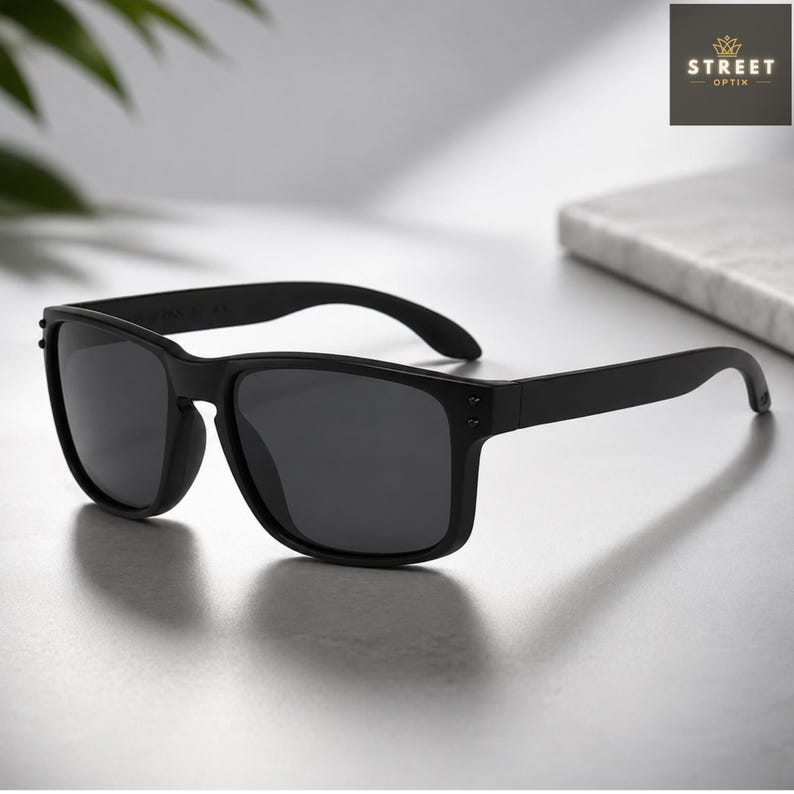 May include: Black sunglasses with a square frame. The sunglasses have dark lenses and a matte finish. The arms of the sunglasses are straight and have a slight curve at the end. The sunglasses are sitting on a light-colored surface.