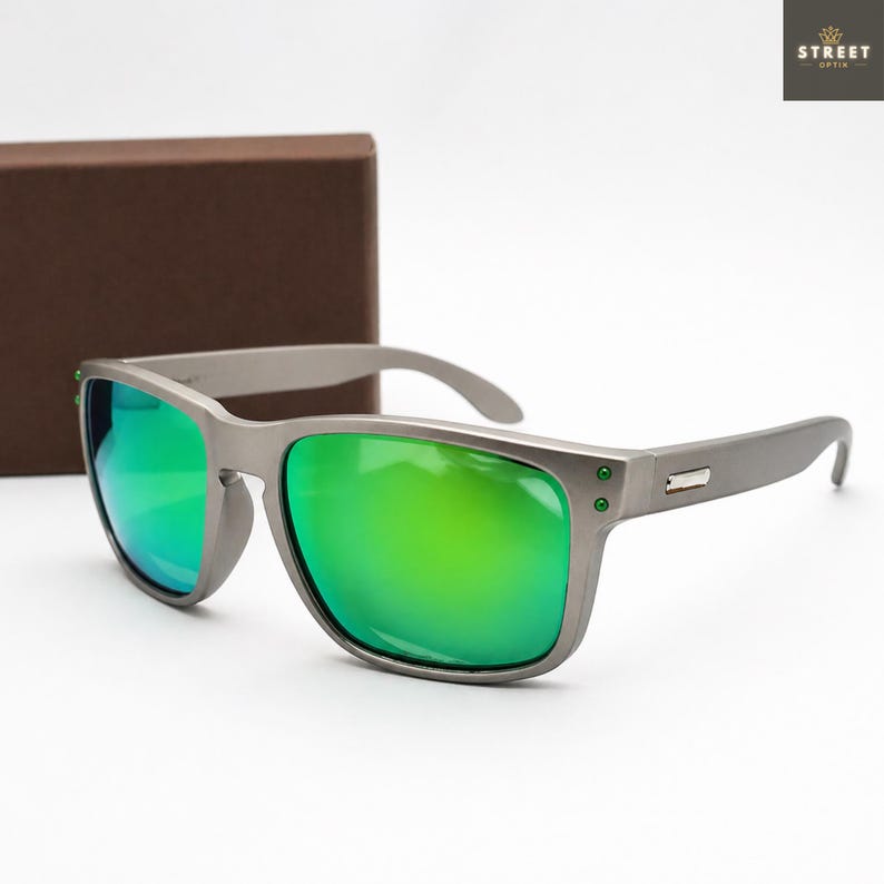 May include: Gray sunglasses with green mirrored lenses. The sunglasses have a rectangular frame with a matte finish and a small metal accent on the temples. The lenses reflect a bright green color. The brand name "STREET OPTIK" is in the top right corner.