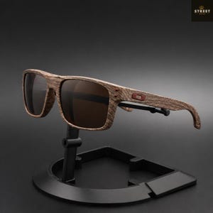 May include: Wooden-framed sunglasses with brown lenses. The frames have a wood grain pattern and a small red accent on the temple. The sunglasses are displayed on a black stand against a dark background. The text "STREET OPTIK" is in the upper right corner.