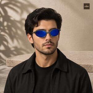 Peut inclure: Lunettes de soleil ovales avec des verres bleus et une monture gris fonc&eacute;. La personne porte une veste noire. L'image a un fond neutre avec le mot "STREET" dans le coin sup&eacute;rieur droit.