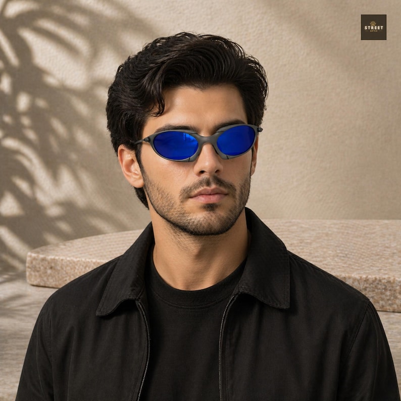 Peut inclure: Lunettes de soleil ovales avec des verres bleus et une monture gris fonc&eacute;. La personne porte une veste noire. L'image a un fond neutre avec le mot "STREET" dans le coin sup&eacute;rieur droit.