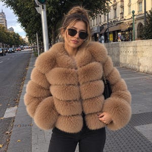 May include: A tan, multi-tiered faux fur jacket with long sleeves and a high collar. The jacket features a horizontal, segmented design with a thick, plush texture. The garment is paired with dark trousers and a black shoulder bag with a gold chain strap.