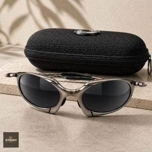 Peut inclure: Des lunettes de soleil &agrave; monture argent&eacute;e et verres fonc&eacute;s sont pos&eacute;es devant un &eacute;tui noir zipp&eacute;. Les lunettes de soleil ont un design &eacute;l&eacute;gant et moderne. L'&eacute;tui a un embl&egrave;me circulaire sur le dessus. Le logo "STREET OPTICS" est en bas &agrave; gauche.