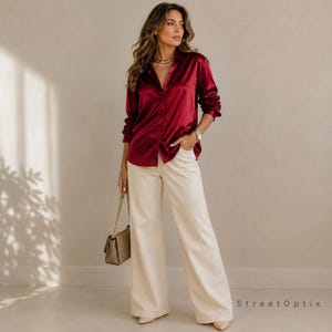 May include: A long-sleeved, button-down shirt in a deep burgundy satin fabric with a collar, paired with high-waisted, wide-leg trousers in a solid cream colour. A small, structured handbag with a gold chain strap is held at the side.