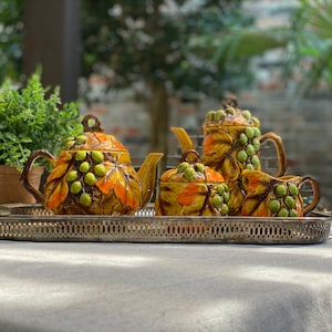 Puede incluir: Juego de té de cerámica vintage con diseño de uvas y hojas en tonos amarillos, naranjas y verdes. El juego incluye una tetera, una crema, un azucarero y una jarrita, todo exhibido en una bandeja plateada.
