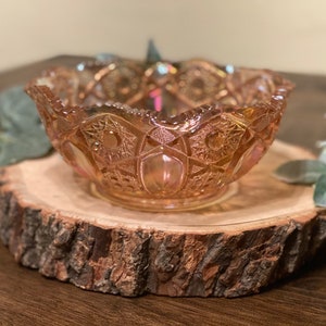 Puede incluir: Un cuenco de vidrio iridiscente vintage con un borde festoneado y un patrón intrincado. El cuenco es de color melocotón pálido con un ligero tono rosado. Está sentado sobre una rodaja de madera.