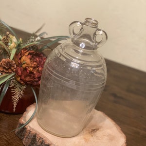 Puede incluir: Una botella de vidrio transparente con dos asas. La botella tiene una superficie texturizada y las palabras "ONE GALLON" son visibles en el lateral. La botella está sentada sobre una rodaja de madera.