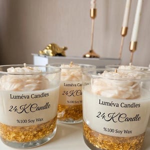 May include: Close-up of several glass candles with a gold leaf base and a white floral design on top. The candles have the text "Luméva Candles 24 K Candle %100 Soy Wax" printed on the glass. Other candles and candle holders are in the background.