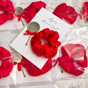 May include: A collection of party favors, each featuring a bright red satin scrunchie and a small round handheld mirror with a gold-toned frame and a red ribbon bow, attached to a white card printed with the text Ayse's Henna Night.