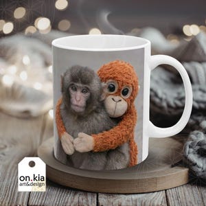 May include: White ceramic mug featuring a heartwarming image of two hugging monkeys. One is a real monkey, the other a plush toy. The mug has a curved handle and the text "on.kia art&design" on a tag.