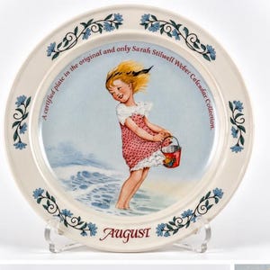 May include: A round ceramic decorative plate featuring a blonde child in a pink polka-dot dress holding a red bucket at the beach. The rim has blue floral vines. Text reads: A certified plate in the original and only Sarah Stilwell Weber Calendar Collection. August.