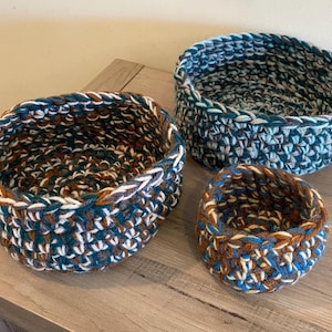 May include: Three round, hand-crocheted baskets of varying sizes. The largest basket features a blend of teal, brown, white, and grey yarn. The medium basket displays shades of blue, teal, and grey, while the smallest basket uses brown, blue, and white.